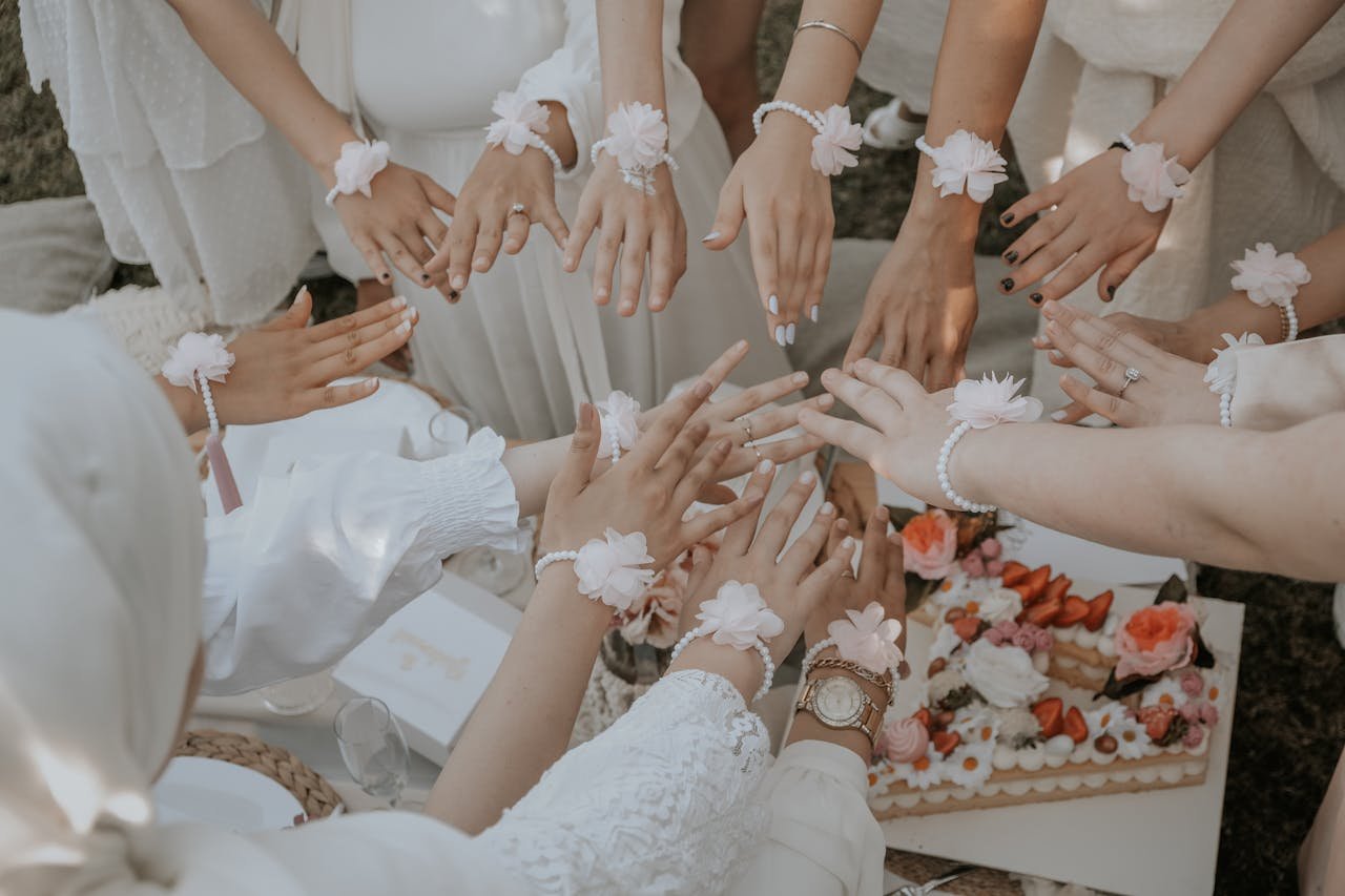 A close-up of women's hands adorned with flower bracelets at a wedding gathering over a decorative cake.